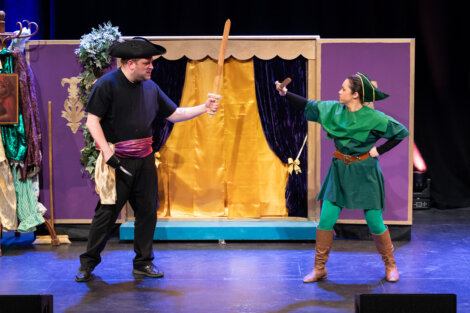 Two actors on stage act out a sword fight, one dressed as a pirate with a wooden sword, the other in a green costume with a small dagger, in front of a purple and gold set.