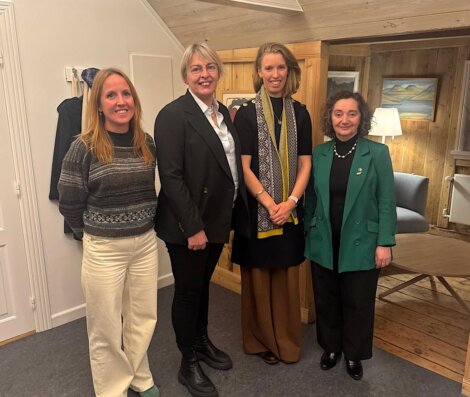 Four women standing indoors, smiling at the camera; they are dressed in business casual attire in a warmly lit room with wood-paneled walls and framed artwork.
