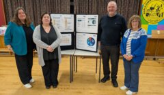 Four adults stand indoors beside a table with charts and graphs displayed on boards, next to a colorful "Scalloway Community Council" banner.