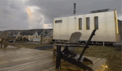 A modern tiny house with vertical siding stands beside a wooden deck with Adirondack chairs, outdoor lights, and a cloudy sky in the background.