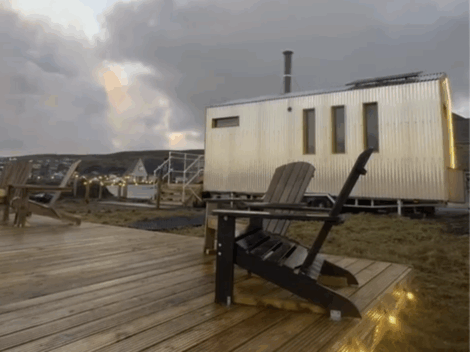 A modern tiny house with vertical siding stands beside a wooden deck with Adirondack chairs, outdoor lights, and a cloudy sky in the background.
