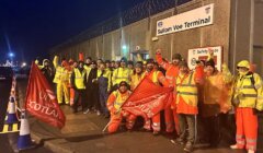A group of workers in high-visibility clothing stand outside Sullom Voe Terminal at night, holding union flags and raising their fists.