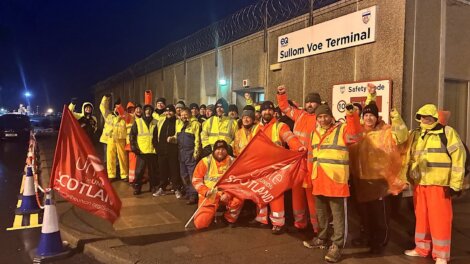 A group of workers in high-visibility clothing stand outside Sullom Voe Terminal at night, holding union flags and raising their fists.