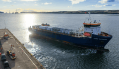 A large cargo ship named EEMS DUBURG is docked at a port, with workers and vehicles visible on the quay under a partly cloudy sky.