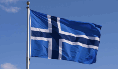 A blue flag with a white and light blue Nordic cross design waves on a flagpole against a clear sky.