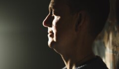 A person in a sweater stands in profile against a wall, illuminated by light from the left, with a neutral expression.