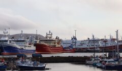 Several large fishing vessels and ferries docked at a harbor, with smaller boats in the foreground and cloudy skies overhead.