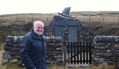 An older man stands in front of a stone memorial with a boat sculpture on top, set in a grassy, hilly landscape behind a wooden gate.