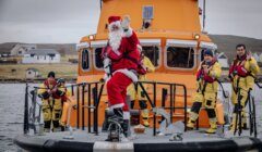 A person dressed as Santa Claus and four crew members in yellow suits stand on the deck of an orange rescue boat, waving towards the camera. Houses and cloudy sky are visible in the background.