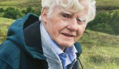 An older man with white hair wearing a green jacket and grey sweater stands outdoors in a grassy, hilly landscape.