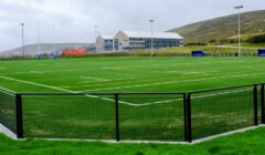An empty outdoor sports field with artificial turf, surrounded by a black fence, with buildings and hills in the background under a cloudy sky.