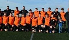 A soccer team in orange jerseys poses together on a field, with coaches and a small child standing on the right side of the group.