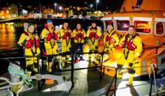 Eight people in yellow rescue suits stand on a lifeboat at night, docked by a waterfront with illuminated buildings in the background.