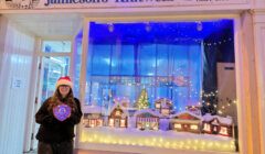 A person in a Santa hat stands outside Jamieson's Knitwear, posing by a festive shop window display with miniature snowy houses and Christmas decorations.