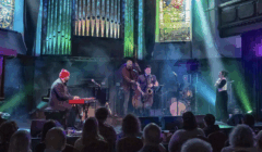 A jazz band performs on stage in a dimly lit venue with stained glass windows, colorful lights, and an audience watching.