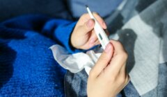 Person in a blue sweater holding a digital thermometer and a tissue, sitting on a plaid blanket.