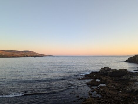 Calm sea at sunset with rocky shore in the foreground and distant hills under a clear sky.