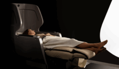 A person reclines in a modern, dark-colored chair with a high backrest, covered by a beige blanket, set against a dark background with a bright curved light.