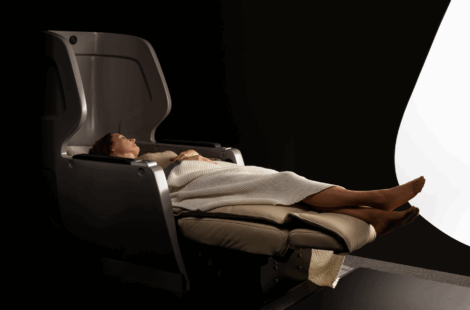 A person reclines in a modern, dark-colored chair with a high backrest, covered by a beige blanket, set against a dark background with a bright curved light.