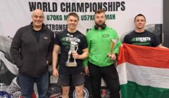 Four men stand in front of a "World Championships" banner; one holds a trophy, another holds a plaque, and the third holds a red, white, and green flag.