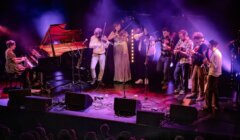 A group of musicians perform on stage with a grand piano, violins, banjo, guitar, and double bass under colorful stage lights.