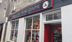 Shopfront of High Level Music Centre with guitars, a drum, and a Santa figure visible through the window; signage displays contact details and a logo above the door.