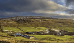 A rural landscape with scattered houses and farm buildings on green fields, set against a gently sloping hill under a cloudy sky.