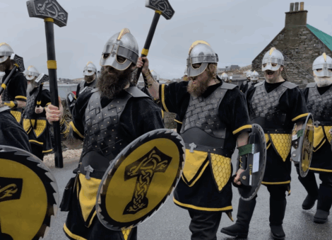 A group of people dressed in Viking-style costumes with helmets, shields, and axes march outdoors on a cloudy day.