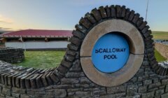A stone wall with a circular blue sign reads "Scalloway Pool." A modern building and grassy area are visible in the background.