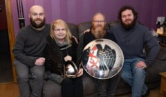 Four people sitting on a couch, one holding a shield with a black bird emblem, another holding a winged helmet, in a living room with purple walls.