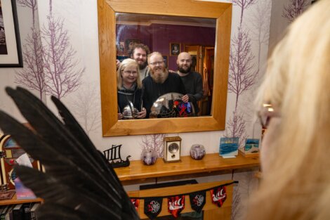 Three people are seen smiling in a mirror, while a person with long blonde hair and black wings stands in the foreground with their back to the camera in a decorated room.