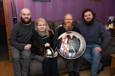 Four people sitting on a couch, one holding a shield with a black bird emblem, another holding a winged helmet, in a living room with purple walls.