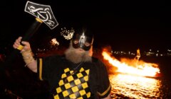 A man in Viking-style armor and helmet holds an axe near a waterfront at night, with a blazing fire on a floating structure in the background.