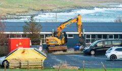 A yellow excavator is parked next to cars and a building near the coast, with waves and grassy hills visible in the background.