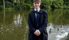 A young person in a graduation gown stands by a pond with ducks and swans, surrounded by trees and greenery.