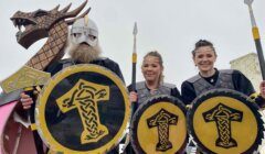 Three people in Viking-inspired costumes holding shields with yellow and black designs, standing in front of a large wooden dragon prop.