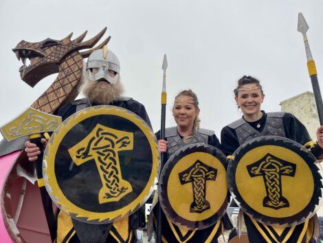 Three people in Viking-inspired costumes holding shields with yellow and black designs, standing in front of a large wooden dragon prop.