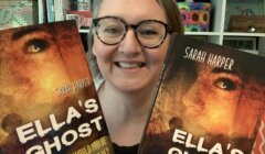 A smiling person with glasses holds up two copies of the book "Ella's Ghost" by Sarah Harper in front of a bookshelf.