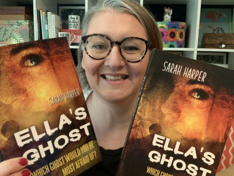 A smiling person with glasses holds up two copies of the book "Ella's Ghost" by Sarah Harper in front of a bookshelf.