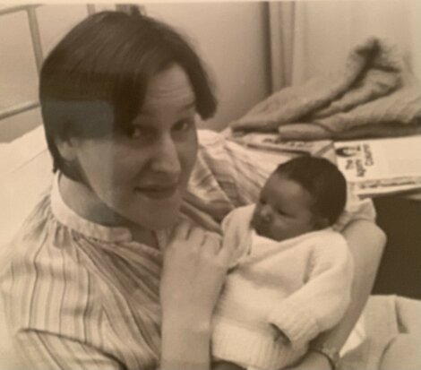 A woman is sitting on a bed and holding a newborn baby wrapped in a white sweater. There are magazines and a blanket in the background.