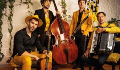 Four men in yellow and black attire pose indoors with musical instruments: a guitar, double bass, clarinet, and accordion. Greenery and artwork decorate the background.