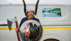 A man wearing a Viking helmet with wings holds an axe and a shield in front of a van with a "Scottish Water" sign.