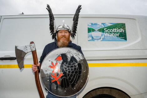 A man wearing a Viking helmet with wings holds an axe and a shield in front of a van with a "Scottish Water" sign.
