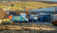 A construction site by the coast with a blue van labeled "Beattie" parked beside fenced-off materials; grassy hills and scattered houses in the background.