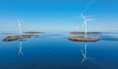 Wind turbines are installed on small rocky islands in a calm blue sea under a clear sky, with reflections visible on the water.