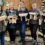 Five women stand indoors in a workshop, each holding a small wooden stool they have made. Various woodworking tools and materials are visible in the background.