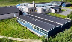 Aerial view of a group of connected school buildings with solar panels on the roofs, surrounded by grass and trees.