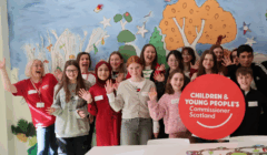 A group of young people and adults stand indoors, smiling and waving at the camera with a sign that reads "Children & Young People's Commissioner Scotland.