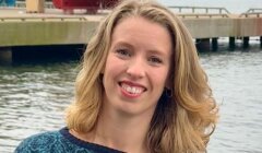 A woman with shoulder-length blonde hair smiles by a waterfront with a dock, boats, and hills in the background.
