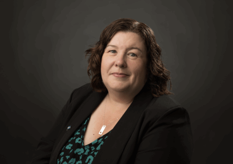 A woman with shoulder-length brown hair wearing a black blazer and patterned top poses in front of a dark, plain background.